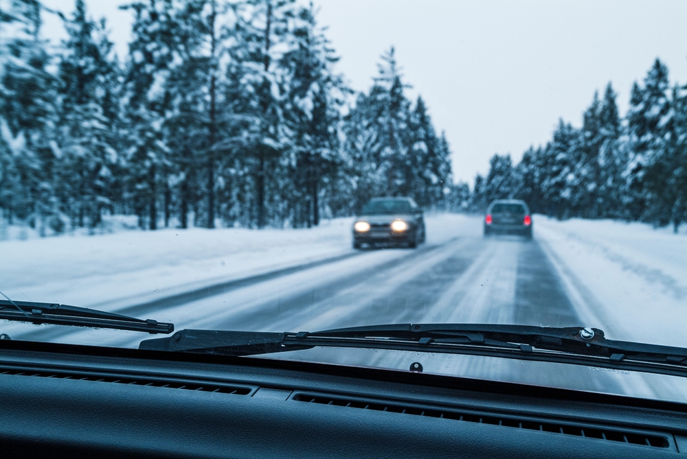 Windshield wiper bladesin focus as vehicles navigate a snowy road surrounded by tall trees on a chilly winter day.